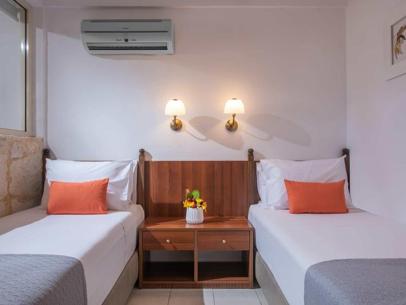 Chambre d'hôtel avec deux lits simples, décoration orange et table de nuit en bois avec fleur.