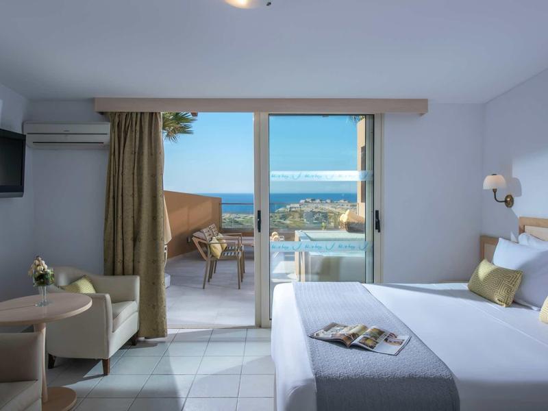Chambre d'hôtel moderne avec balcon et vue sur la mer, lit confortable et coin salon.