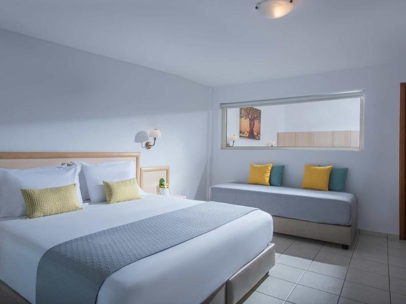 Chambre d'hôtel moderne avec grand lit double et coin salon près de la fenêtre avec coussins colorés.