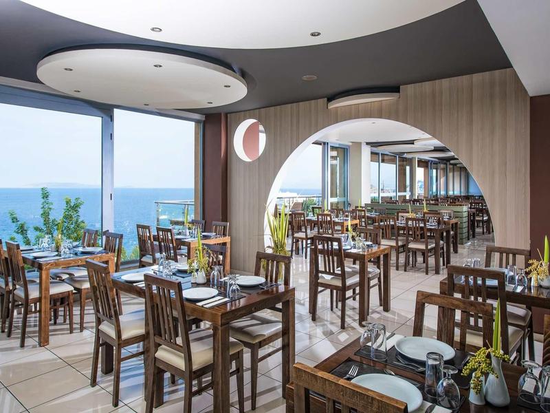 Restaurant moderne avec tables et chaises en bois, vue sur la mer à travers de grandes fenêtres.