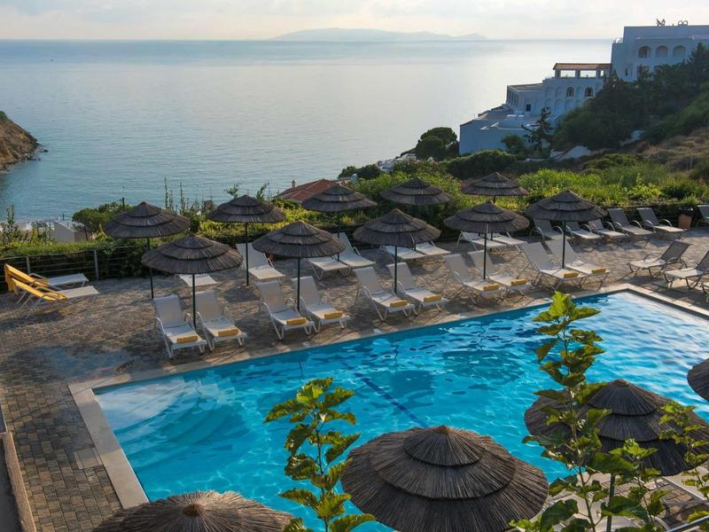 Piscine avec parasols surplombant la mer et des bâtiments proches au coucher du soleil.