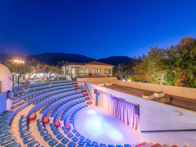 Teatro all'aperto con sedili blu e palco illuminato sotto un cielo limpido al crepuscolo.