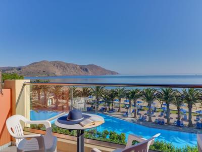 Balcone con tavolo e sedie con vista su piscina, palme e mare sotto un cielo limpido.
