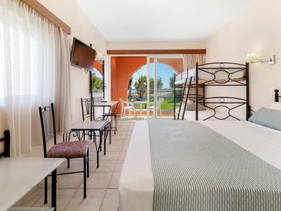 Světý hotelový pokoj s manželskou postelí, psacím stolem, židlemi a terasou s výhledem na zahradu a moře.