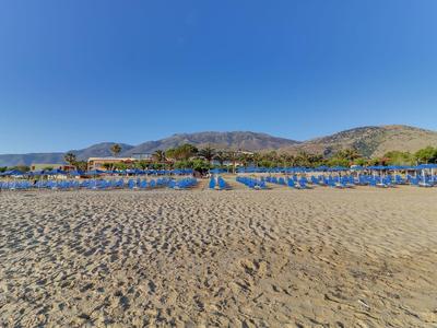 Široká písečná pláž s řadami modrých slunečníků a horami v pozadí za jasné oblohy.