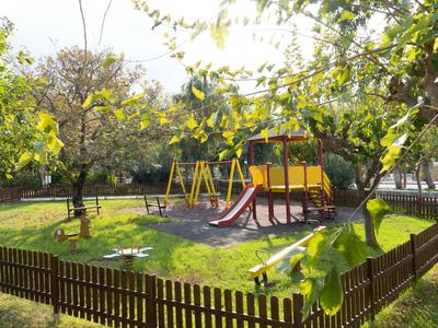 Dětské hřiště s klouzačkou a houpačkami, obklopené dřevěným plotem a stromy