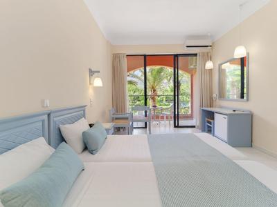 Světly hotelový pokoj se dvěma postelemi, balkonem a výhledem na moře.