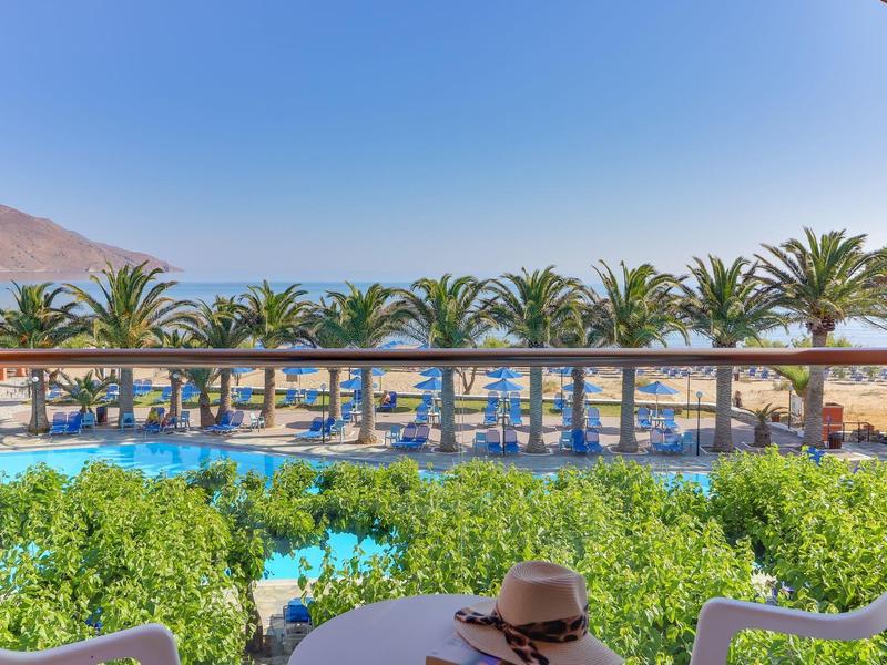 Výhled na hotelový bazén s palmami a lehátky na pláži za jasné oblohy.