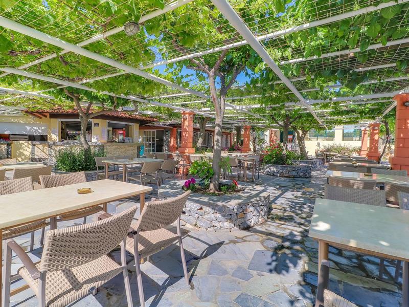 Slunná terasa s stolky a židlemi pod zelenou pergolou v hotelu.