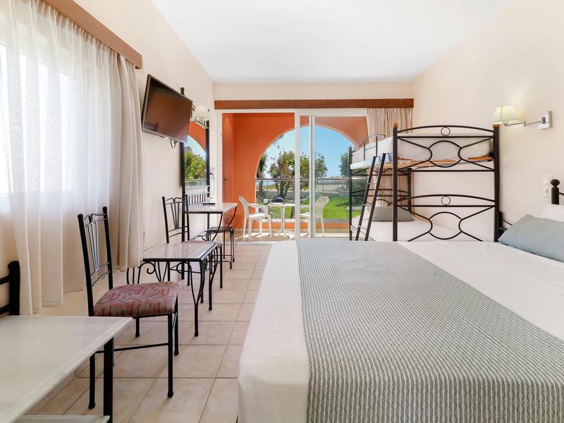 Světý hotelový pokoj s manželskou postelí, psacím stolem, židlemi a terasou s výhledem na zahradu a moře.