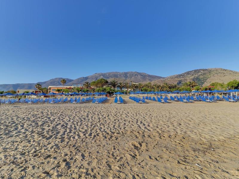 Široká písečná pláž s řadami modrých slunečníků a horami v pozadí za jasné oblohy.