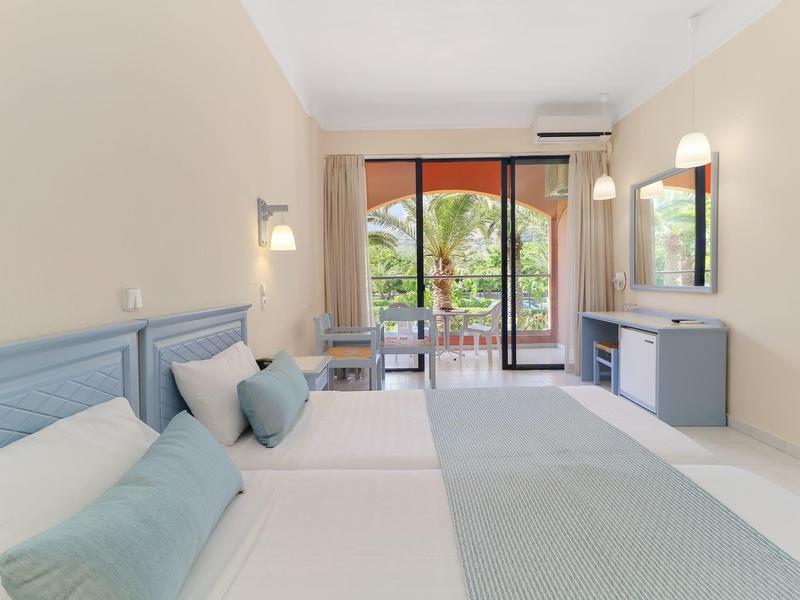 Světly hotelový pokoj se dvěma postelemi, balkonem a výhledem na moře.