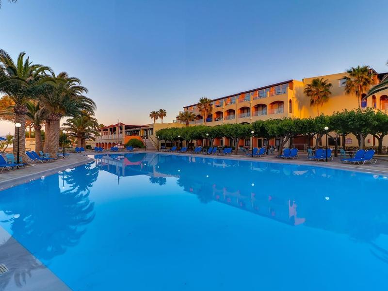 Velký venkovní bazén s lehátky před hotelovým komplexem v středomořském stylu za jasné oblohy