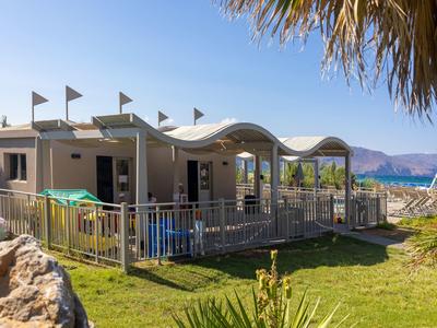 Kleines Strandhaus mit wellenförmigem Dach, draußen Zäune, Palmen und grüner Rasen am Meer.