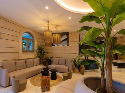 Sala moderna de hotel con paredes de madera, sofás, plantas y mesa de centro redonda bajo luz cálida.