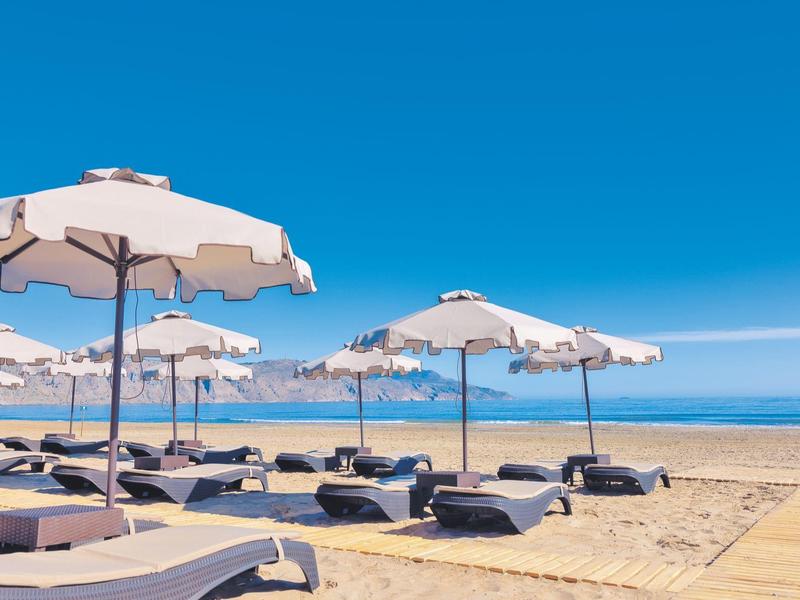 Playa con sombrillas blancas y tumbonas bajo un cielo azul claro junto al mar.