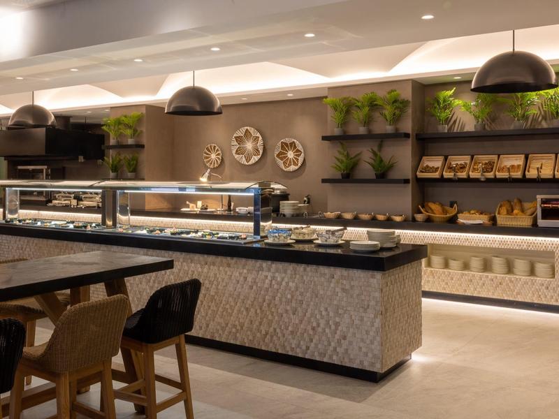 Estación de buffet moderna en hotel con iluminación, plantas y asientos en tonos cálidos.