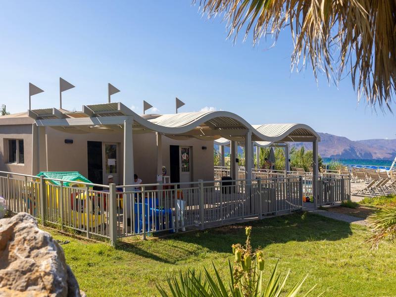 Casa pequeña con techo ondulado y terraza junto al mar, rodeada de vegetación y rocas.