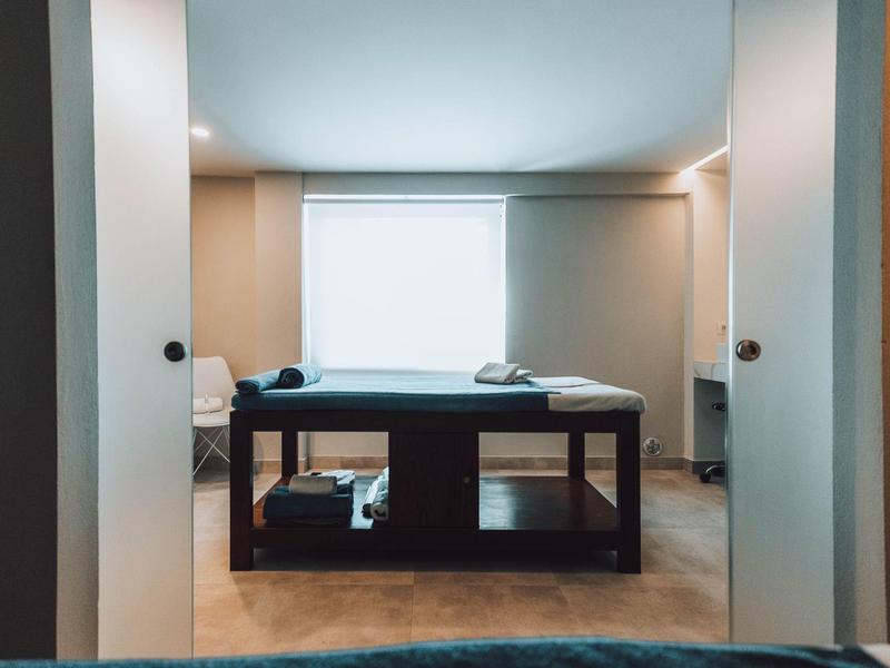 Sala de masajes con diseño moderno, cama de masaje centrada frente a ventana luminosa.