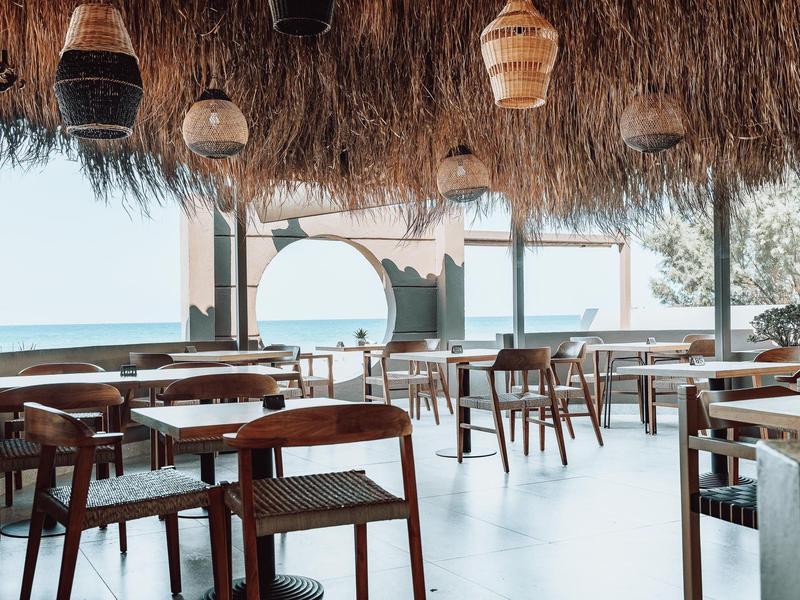 Restaurante abierto con muebles de madera y vista al mar bajo un techo de paja.