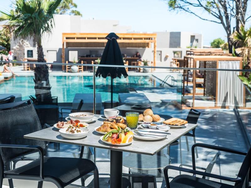 Mesa con desayuno y bebidas junto a una piscina en un hotel con palmeras y edificios al fondo.