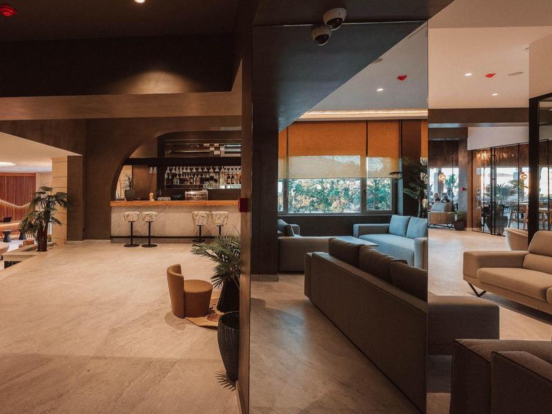 Área moderna del lobby del hotel con zonas de estar, muebles de madera y vista a un bar al fondo.