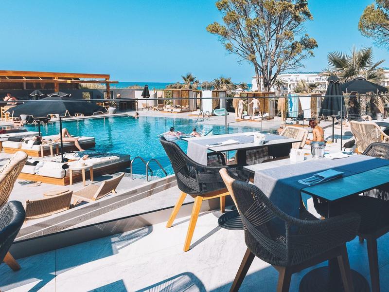 Área exterior moderna de hotel con piscina, mesas, sillas y vista al mar en plena luz del día.