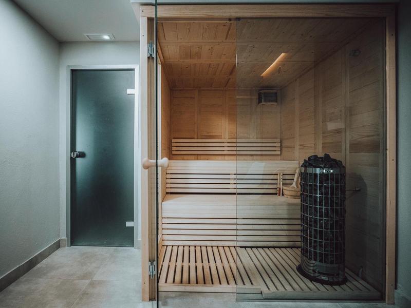 Sauna moderna con pared de vidrio, bancos de madera y horno eléctrico en un área de bienestar.