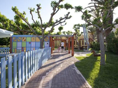 Pfade und bunte Zäune entlang eines sonnigen Hotelgartens mit Bäumen und Menschen.