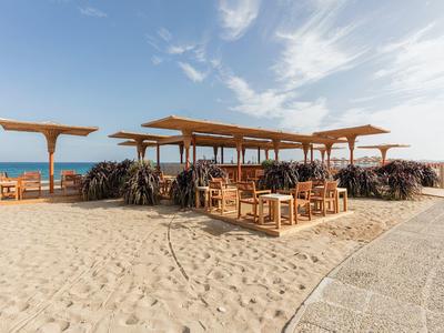 Offener Strandbereich mit Holzmöbeln und Sonnendächern auf Sand bei klarem Himmel.