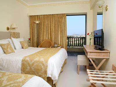 Helles Hotelzimmer mit zwei Betten, goldfarbenen Vorhängen und Balkon mit Meerblick.