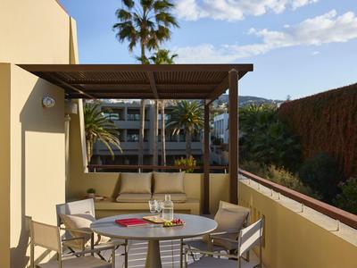 Terraza con mesa, sillas y pérgola, rodeada de edificios de hotel y palmeras bajo un cielo azul.