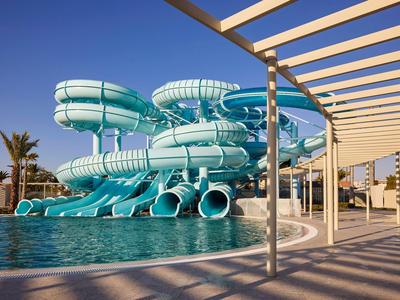 Toboganes de agua azules sobre una piscina en un lugar de vacaciones soleado con pérgola y palmeras.