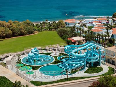Parque acuático con varios toboganes azules junto a un campo de golf y edificios de hotel junto al mar.