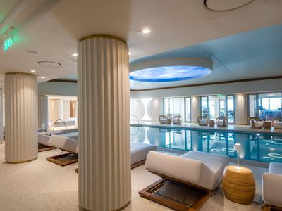 Zona de piscina interior con columnas, tumbonas cómodas e iluminación indirecta en un hotel.