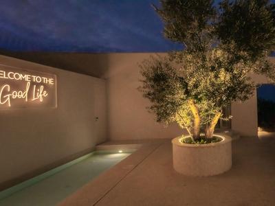 Área exterior moderna con árbol iluminado y proyección en la pared "Welcome to the Good life" de noche.