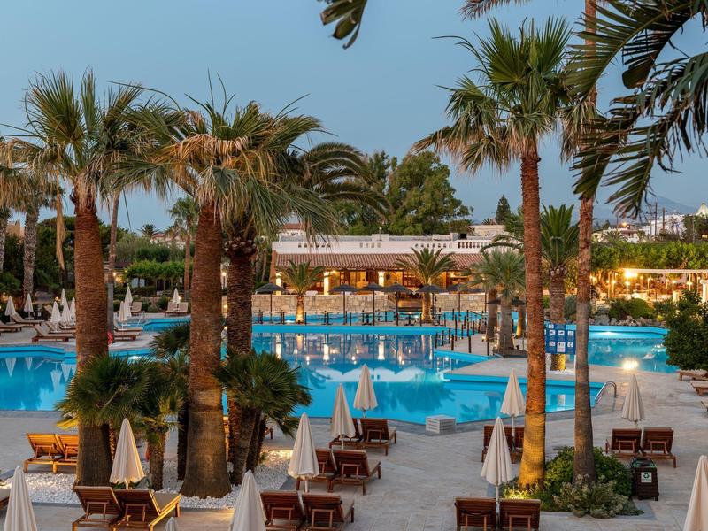 Großer Hotelpool mit Palmen, Liegestühlen und Sonnenschirmen bei klarem Himmel.