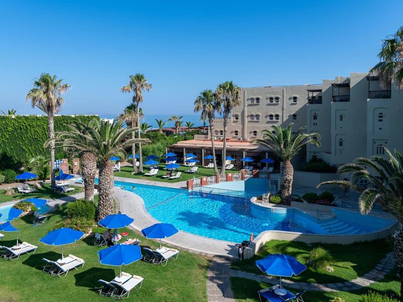 Hotelpool mit Liegestühlen, Palmen und umliegendem Garten unter blauem Himmel.
