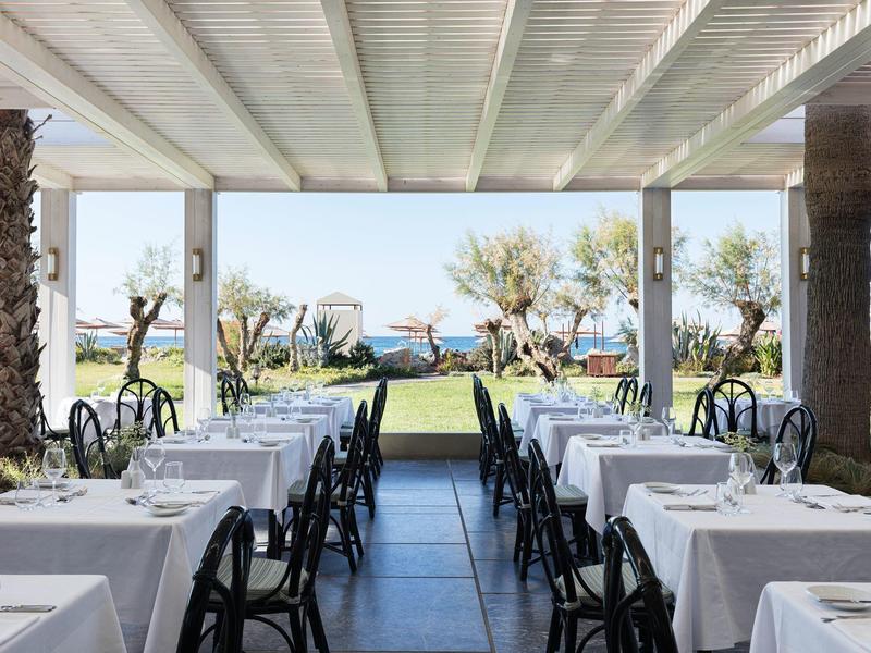 Restaurant mit weißen Tischtüchern und Blick auf den Garten und das Meer im Hintergrund.