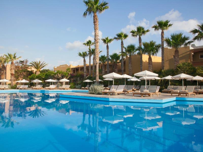 Ein großer blauer Pool mit Palmen und Sonnenschirmen im Hotelresort bei strahlendem Wetter.