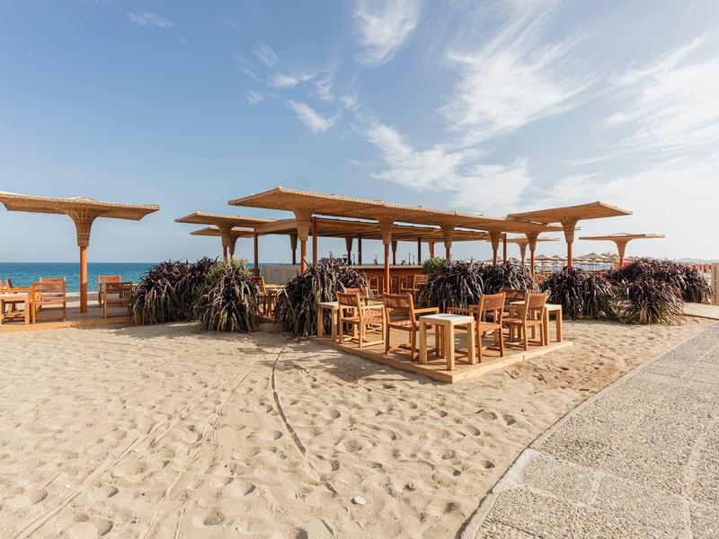 Offener Strandbereich mit Holzmöbeln und Sonnendächern auf Sand bei klarem Himmel.