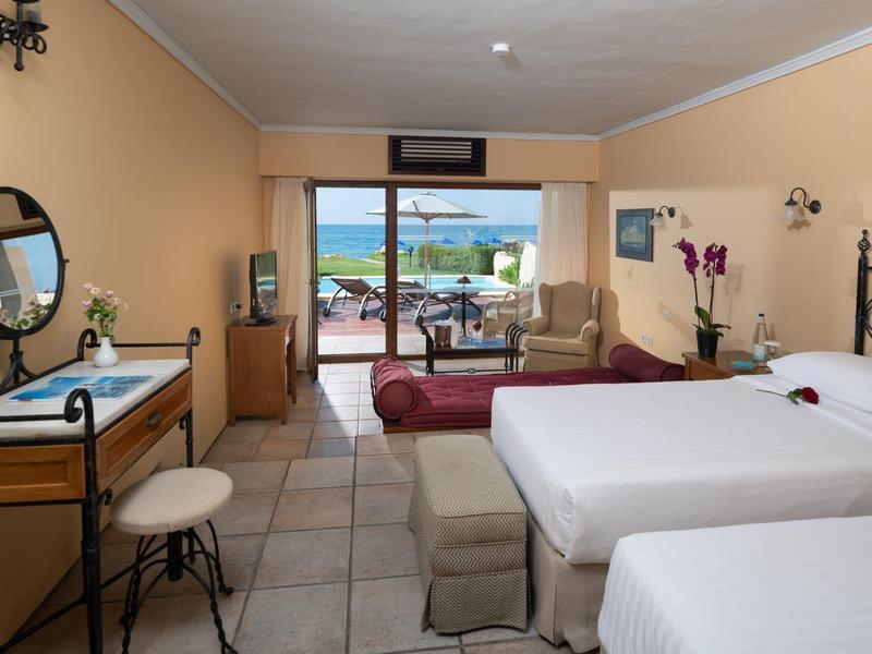 Hotelzimmer mit zwei Betten, Schreibtisch, Sessel und Blick auf Terrasse und Meer.