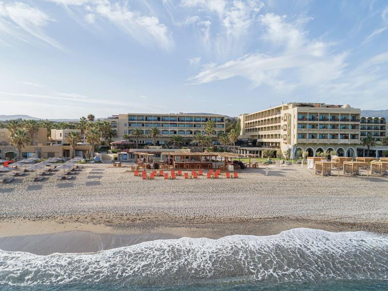 Ein Strandhotel mit Liegestühlen und Tischen direkt am Sandstrand unter blauem Himmel.