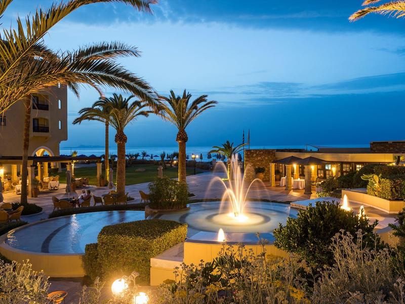 Abendliche Hotelanlage mit beleuchtetem Brunnen, Pools und Palmen unter blauem Himmel.