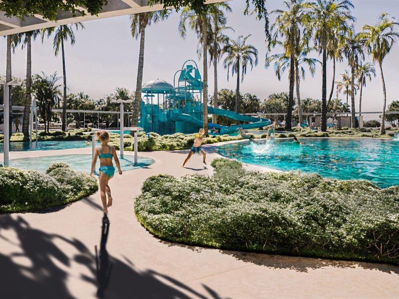 Außenpool mit Wasserrutschen, Palmen und Sonnenschirmen in tropischer Hotelanlage.