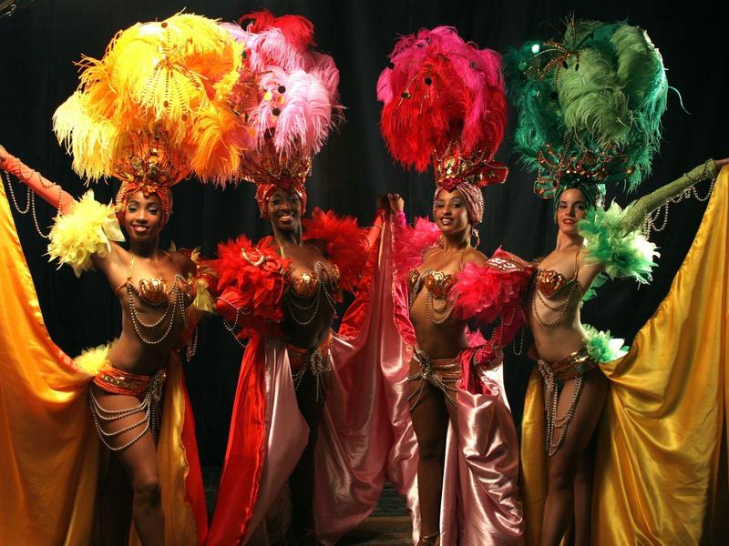 Vier Tänzerinnen in farbenfrohen Kostümen mit großen Federkopfschmuck auf dunklem Hintergrund.