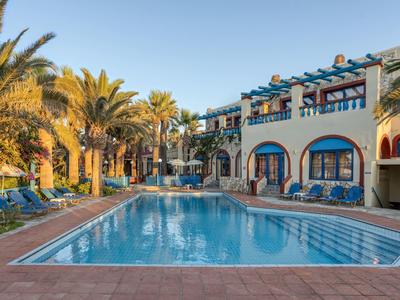 Hotelzwembad met ligstoelen en palmbomen onder een heldere lucht naast een Mediterraan gebouw.