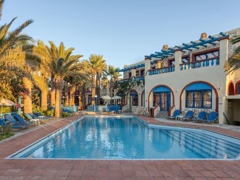 Hotel con piscina e palme sotto un cielo azzurro, circondato da edifici bianchi.