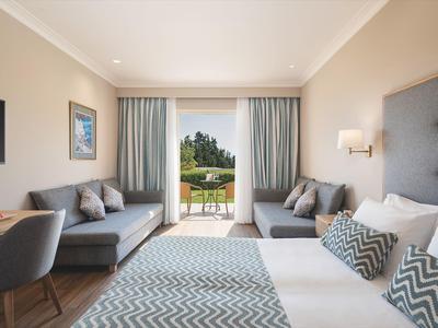 Moderní hotelový pokoj s manželskou postelí, pohovkami a balkonem s výhledem na zeleň.