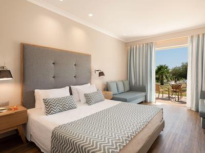 Světly hotelový pokoj s manželskou postelí, pohovkou, lampičkami a přístupem na balkon s výhledem.